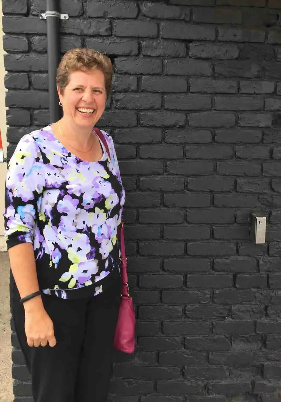 Joanne, 51, smiling outdoors in front of a dark brick wall